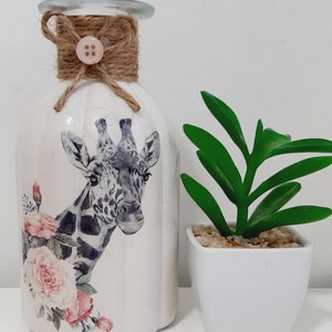 May include: A white ceramic vase with a giraffe and pink roses design. The vase has a brown twine bow with a button on top. A small white planter with a green succulent plant is next to the vase.