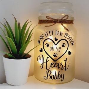 May include: A yellow glass jar with a string tied around the top. The jar has a black and white design that says "You left paw prints on my heart Bobby".