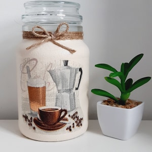 May include: A white glass jar with a brown twine bow and a coffee themed design. The design features a coffee pot, a cup of coffee, and coffee beans. The jar is sitting on a white surface.