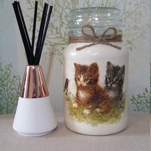 May include: A white glass jar with a brown twine bow and a printed image of two kittens and a bird. The jar is sitting on a beige surface next to a white diffuser with a copper top.