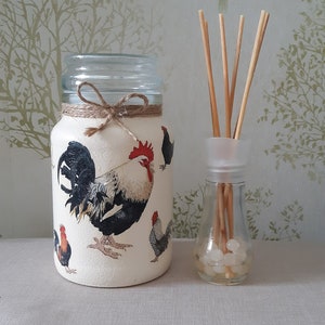 May include: A white glass jar with a rooster and chickens design, tied with twine, and filled with clear liquid. A clear glass bottle with a white lid and wooden sticks is next to the jar.
