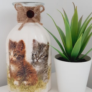 May include: A white ceramic vase with a brown twine bow and a brown button. The vase features a picture of two kittens. The vase is on a white surface next to a white pot with a green plant.