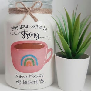 May include: A white jar with a glass lid, decorated with a pink coffee cup design and a rainbow. The jar has the text "May your coffee be strong & your Monday be short." A small potted plant is next to the jar.
