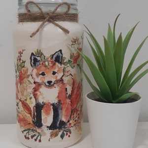 May include: A white glass jar with a brown twine bow and a fox illustration. The jar features a watercolour design of a fox wearing a floral wreath. The fox is surrounded by flowers and leaves in shades of orange, red, yellow, and green.