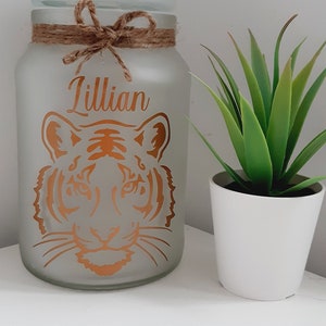 May include: A frosted glass jar with a brown twine bow and the name "Lillian" written in gold lettering. A tiger face is also printed in gold on the jar. A small potted green plant is in the background.