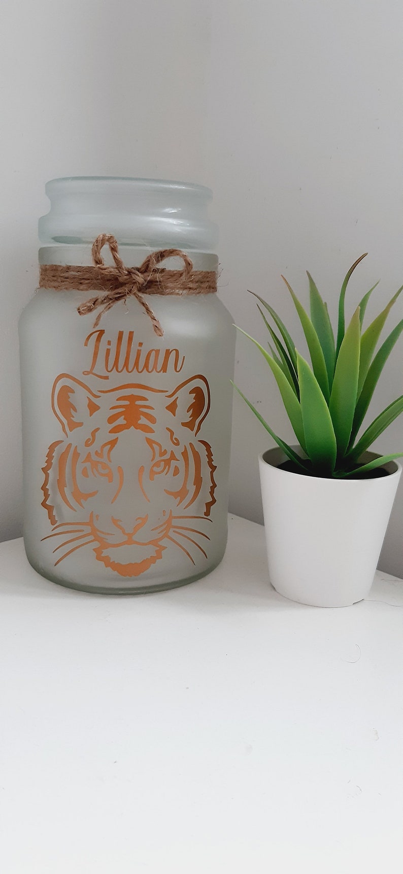 May include: A frosted glass jar with a brown twine bow and the name "Lillian" written in gold lettering. A tiger face is also printed in gold on the jar. A small potted green plant is in the background.