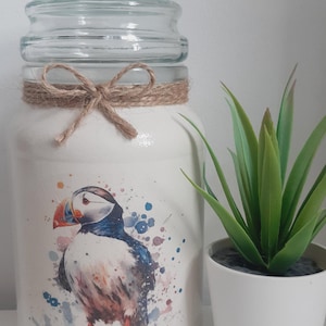 May include: A glass jar with a lid, decorated with a watercolour puffin design and twine. The puffin has a red and orange beak, and is surrounded by blue and purple paint splatters. A small potted plant sits beside the jar.