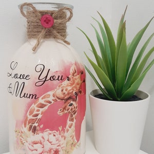May include: White ceramic vase with a pink and brown giraffe design. The vase has a brown twine bow with a pink button on top. The vase says "Love You Mum" on the front. The vase is sitting on a white surface next to a small green plant in a white pot.