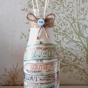 May include: A decorative glass bottle with a beach theme, featuring text like "Beach Cottage" and "Havana 105." The bottle is adorned with twine, a blue button, and wooden reeds for a diffuser.