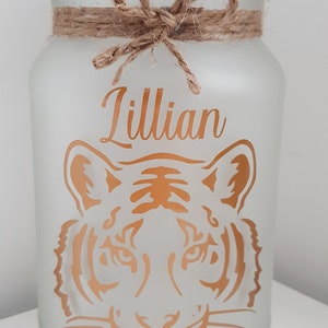 May include: A frosted glass jar with a brown twine bow and a gold tiger face design. The name "Lillian" is written in gold lettering on the jar.