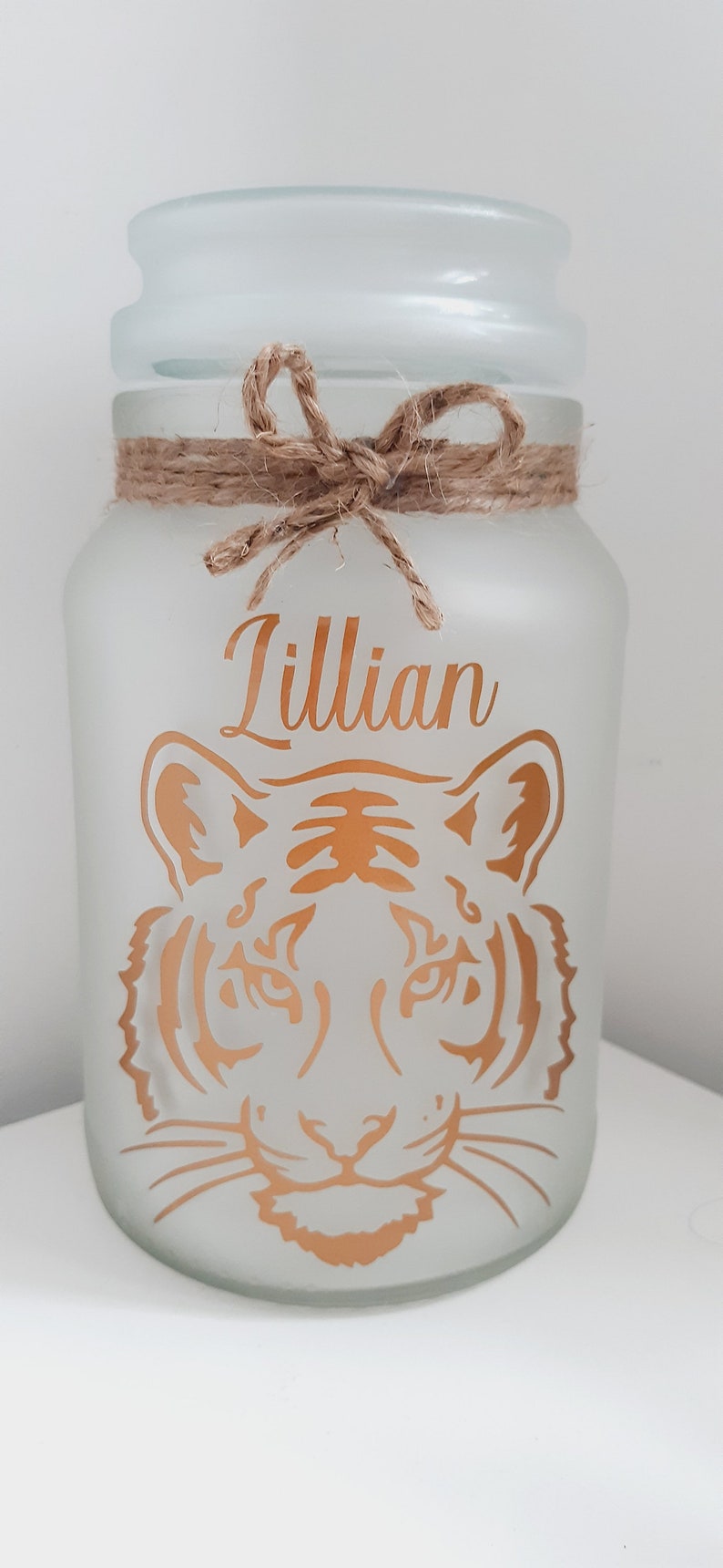 May include: A frosted glass jar with a brown twine bow and a gold tiger face design. The name "Lillian" is written in gold lettering on the jar.