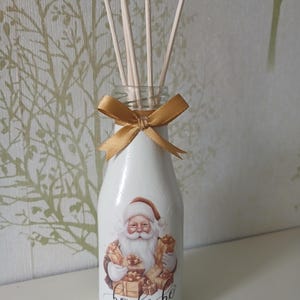 May include: A white glass bottle diffuser featuring a graphic of Santa Claus holding wrapped gifts with the text ho ho ho printed below. The bottle neck is decorated with a gold satin ribbon bow and contains four light-coloured wooden reed sticks.