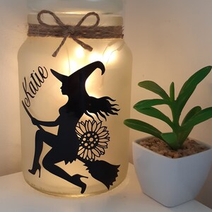 May include: A frosted glass jar with a string bow and a silhouette of a witch flying on a broomstick with a sunflower behind her. The jar has a string of lights inside and the name "Katie" is written on the side.
