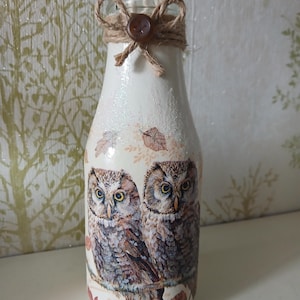 May include: A glass bottle painted in cream with a glittery finish, featuring a detailed illustration of two owls perched on a branch. The neck of the bottle is wrapped in brown twine tied into a bow with a small brown button at the centre.