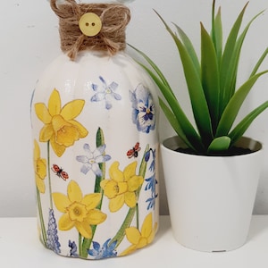 May include: A white ceramic vase with a floral design featuring yellow daffodils, blue forget-me-nots, and white flowers. The vase has a brown twine bow with a yellow button on top.