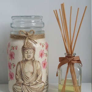 K&ouml;nnte beinhalten: Ein Glasgef&auml;&szlig; mit wei&szlig;em Hintergrund und einem gemalten Bild eines meditierenden Buddha mit rosa Blumen. Das Gef&auml;&szlig; hat eine braune Kordelschleife um den oberen Rand. Neben dem Gef&auml;&szlig; steht eine durchsichtige Glasflasche mit einer braunen Schleife und Holzst&auml;bchen.