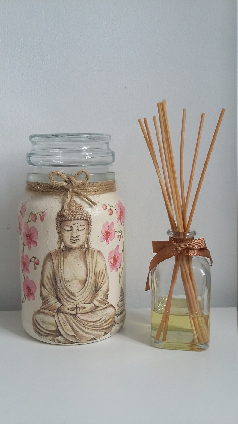 K&ouml;nnte beinhalten: Ein Glasgef&auml;&szlig; mit wei&szlig;em Hintergrund und einem gemalten Bild eines meditierenden Buddha mit rosa Blumen. Das Gef&auml;&szlig; hat eine braune Kordelschleife um den oberen Rand. Neben dem Gef&auml;&szlig; steht eine durchsichtige Glasflasche mit einer braunen Schleife und Holzst&auml;bchen.