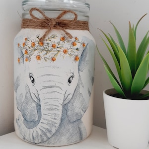 May include: A glass jar with a light pink background and a hand-painted illustration of a grey elephant wearing a flower crown. The jar is tied with a brown twine bow.