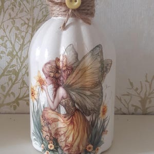 May include: White decorative vase featuring a fairy illustration. The vase has a clear glass top, adorned with twine and a yellow button. The fairy has yellow and brown wings, kneeling near yellow flowers.