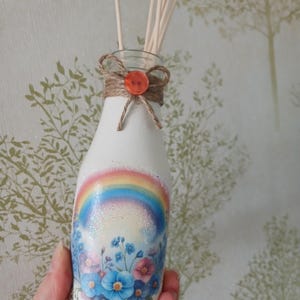May include: White glass diffuser bottle featuring a rainbow and floral design. The bottle is decorated with a rainbow, glitter, and blue and pink flowers. It has a twine bow and an orange button. Wooden diffuser sticks are inserted into the bottle.