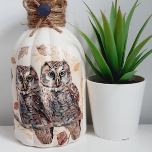 May include: A white ceramic vase with a brown twine bow and a black button. The vase features a design of two owls and autumn leaves. A small green plant in a white pot sits beside the vase.