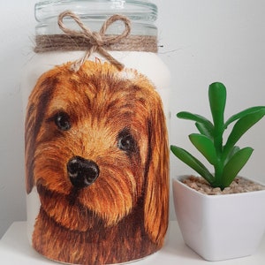 May include: A glass jar with a brown dog illustration on the front. The jar has a brown twine bow around the top. The jar is sitting on a white surface next to a small potted plant.