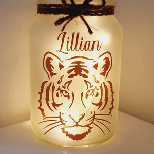 May include: A white frosted glass jar with a brown twine bow and a copper tiger design. The jar is lit from within and the name "Lillian" is written on the front.