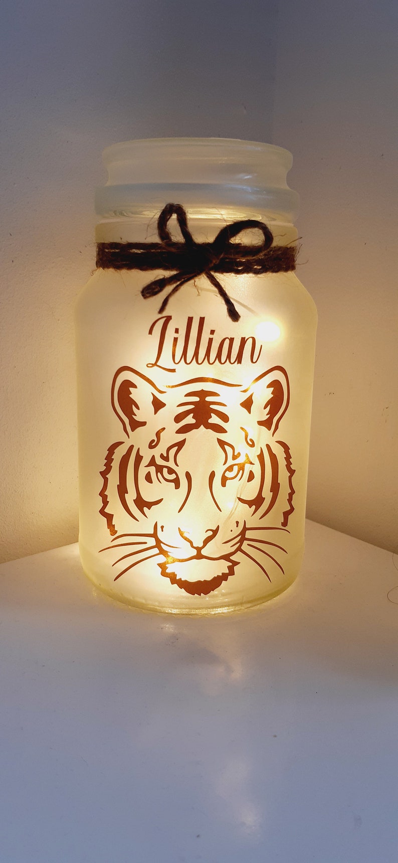May include: A white frosted glass jar with a brown twine bow and a copper tiger design. The jar is lit from within and the name "Lillian" is written on the front.