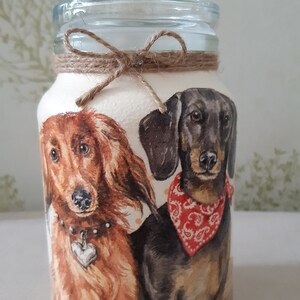 K&ouml;nnte beinhalten: Ein Glasgef&auml;&szlig; mit einer braunen Kordelschleife und einem wei&szlig;en Hintergrund, auf dem zwei Dackel abgebildet sind. Ein Hund ist braun und der andere ist schwarz mit einem roten Halstuch.