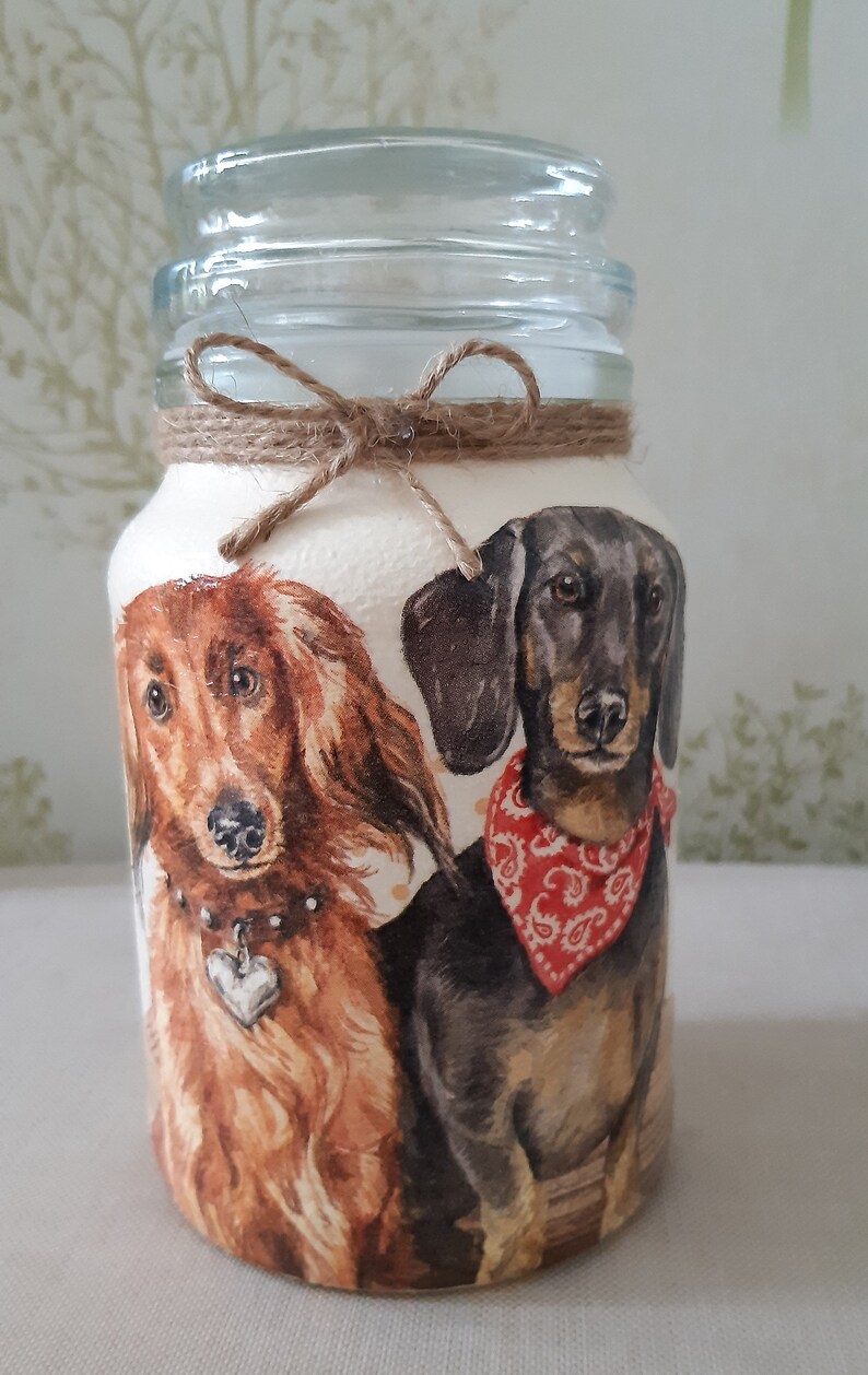 K&ouml;nnte beinhalten: Ein Glasgef&auml;&szlig; mit einer braunen Kordelschleife und einem wei&szlig;en Hintergrund, auf dem zwei Dackel abgebildet sind. Ein Hund ist braun und der andere ist schwarz mit einem roten Halstuch.