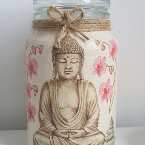 K&ouml;nnte beinhalten: Ein Glasgef&auml;&szlig; mit einer braunen Kordelschleife und wei&szlig;em Hintergrund. Das Gef&auml;&szlig; zeigt ein gemaltes Bild eines meditierenden Buddha mit rosa Orchideenbl&uuml;ten.