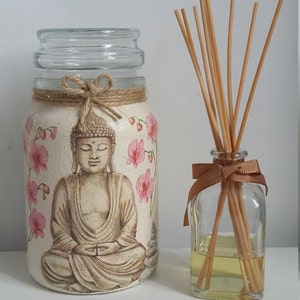 K&ouml;nnte beinhalten: Ein Glasgef&auml;&szlig; mit wei&szlig;em Hintergrund und einem gemalten Bild eines meditierenden Buddha, umgeben von rosa Blumen. Das Gef&auml;&szlig; hat eine braune Kordelschleife und einen durchsichtigen Glasdeckel. Eine durchsichtige Glasflasche mit braunem Band und Holzst&auml;bchen steht neben dem Gef&auml;&szlig;.