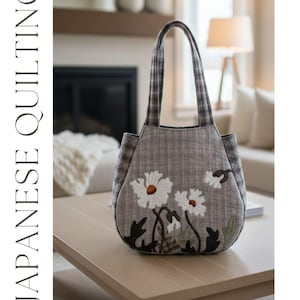 Peut inclure: Un sac fourre-tout matelassé gris avec des appliqués de marguerites blanches et des tiges et feuilles noires. Le sac a une poignée et des panneaux latéraux à carreaux. Les mots "JAPANESE QUILTING" sont imprimés verticalement à gauche. L'adresse du site web est imprimée en bas.