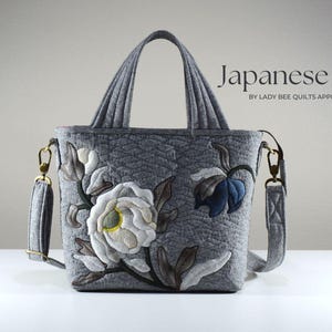 May include: Gray quilted Japanese bag with floral applique. The bag features a white and yellow flower, a blue flower, and brown leaves. It has a top handle and a detachable shoulder strap. The text "Japanese Bag" is displayed on the bag.