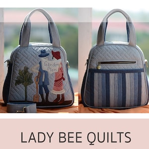 May include: A blue quilted handbag with a floral design and the text "Garden Plan". The bag has a top handle and a shoulder strap. The bag is 28 cm long, 25 cm wide, and 12 cm high.