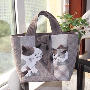May include: A grey quilted tote bag with two embroidered cats on the front. The cats are white with brown spots and have black eyes. The bag has two brown handles.