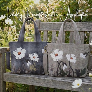 May include: Two tote bags with daisy applique designs sit on a wooden bench. One bag is dark gray, the other is tan. Both have white daisies with brown centers and green leaves. The bags have handles and shoulder straps.