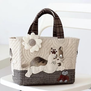 以下が含まれることがあります： ベージュのキルティングトートバッグ。遊び心のある猫のアップリケが施されています。茶色と白の花と、茶色と白のストライプのハンドルが付いています。漫画の猫と赤い車もバッグにアップリケされています。