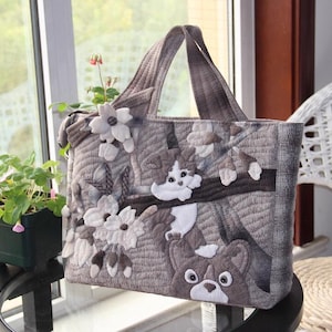 May include: A gray quilted tote bag with a floral design and two embroidered cats. The bag has a brown handle and a zipper closure.