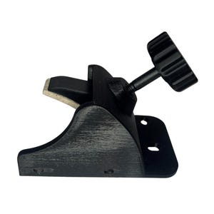 May include: A black clamp with a textured finish, featuring a screw and a knurled knob for adjustment. The clamp has a curved base and a padded contact surface. The design includes two mounting holes.