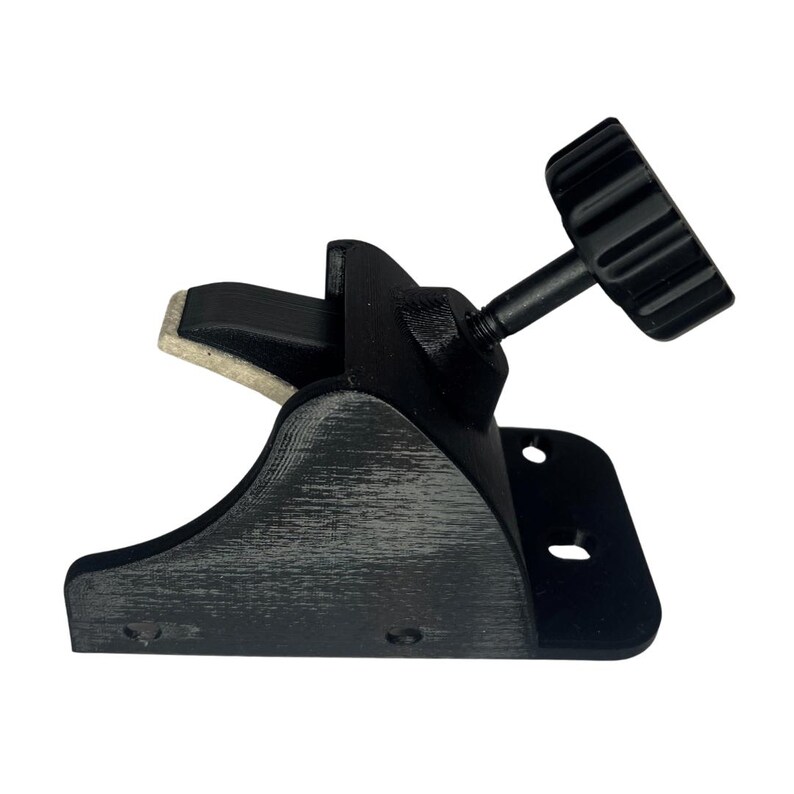 May include: A black clamp with a textured finish, featuring a screw and a knurled knob for adjustment. The clamp has a curved base and a padded contact surface. The design includes two mounting holes.