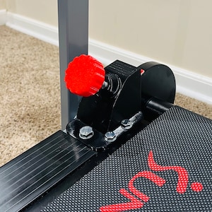 May include: Close-up of a black exercise machine component with a red adjustment knob. The machine has a textured black surface and the word "Sunny" in red. The background includes a wall and a carpeted floor.