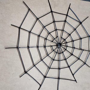 May include: A black spider web decoration constructed from thin, rigid material. The design features a central point with radiating spokes connected by concentric, curved segments, forming a symmetrical, multi-layered web pattern.