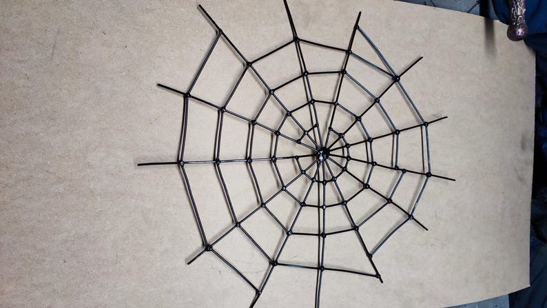 May include: A black spider web decoration constructed from thin, rigid material. The design features a central point with radiating spokes connected by concentric, curved segments, forming a symmetrical, multi-layered web pattern.