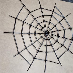 May include: A black spider web decoration constructed from thin, rigid material. The design features a central hub with radiating spokes connected by concentric circular segments, forming a geometric web pattern with a symmetrical structure.