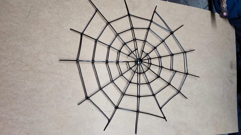 May include: A black spider web decoration constructed from thin, rigid material. The design features a central hub with radiating spokes connected by concentric circular segments, forming a geometric web pattern with a symmetrical structure.