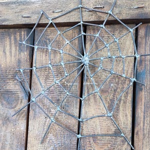 May include: A spider web sculpture constructed from thin, dark grey metal rods joined at intersecting points with visible weld marks. The web features a central hub with radiating spokes connected by concentric, angular segments of metal wire.