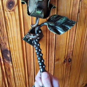 May include: A handcrafted metal rose sculpture with a dark green and bronze finish. The rose has detailed petals and leaves, and is held by a hand. The sculpture is a decorative item.