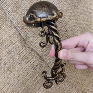 Op de afbeelding: Een handgemaakte metalen deurklink in bronskleur. De klink heeft een paddenstoelvormige bovenkant met textuurdetails en een gedraaide steel. Decoratieve krullen en wervelingen sieren de steel en de basis.