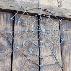 May include: A spider web sculpture crafted from dark grey metal rods. The design features a central hub with radiating spokes connected by concentric circular segments, all joined together with visible welded joints at each intersection point.